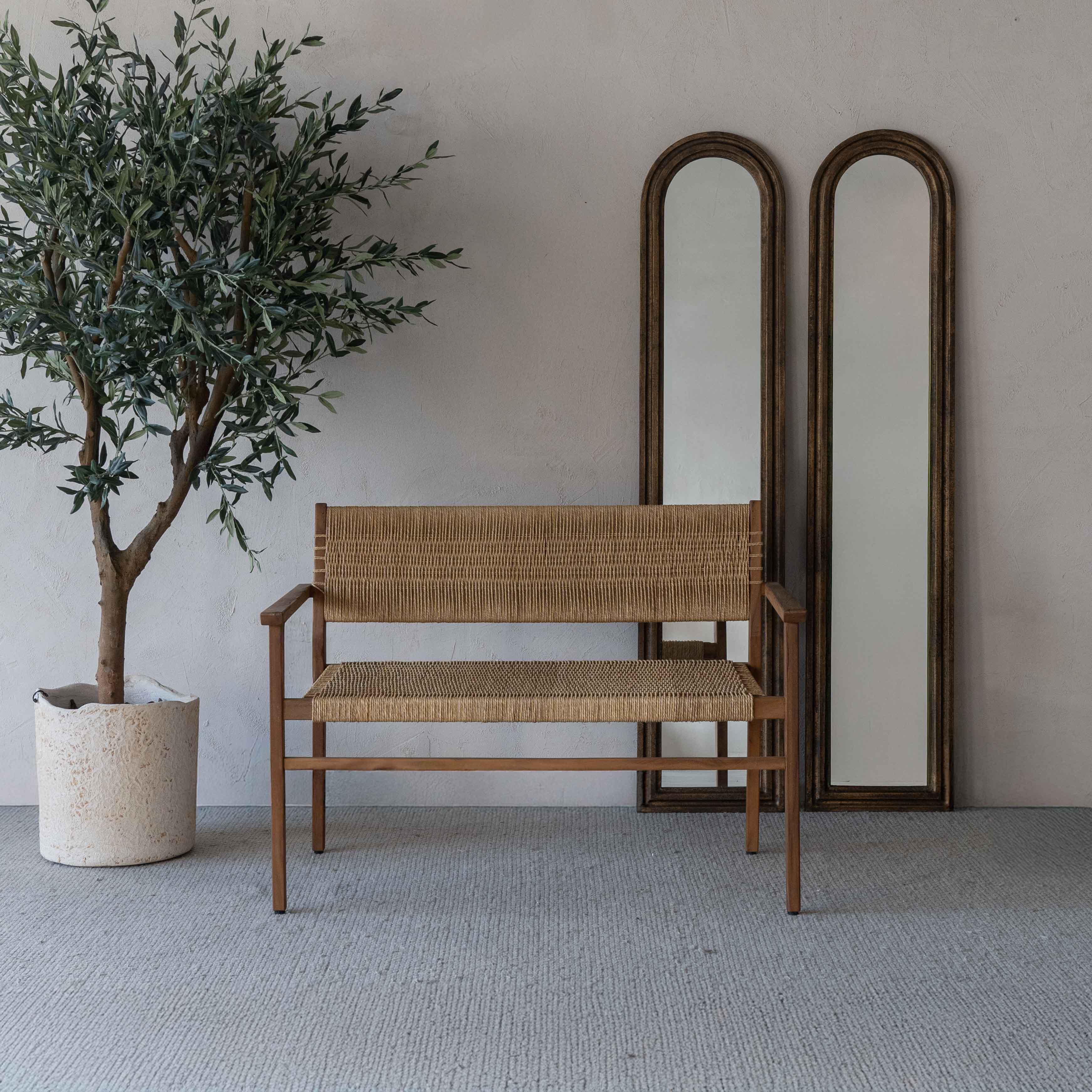 Woven bench next to a potted plant and two tall mirrors against a plain wall.