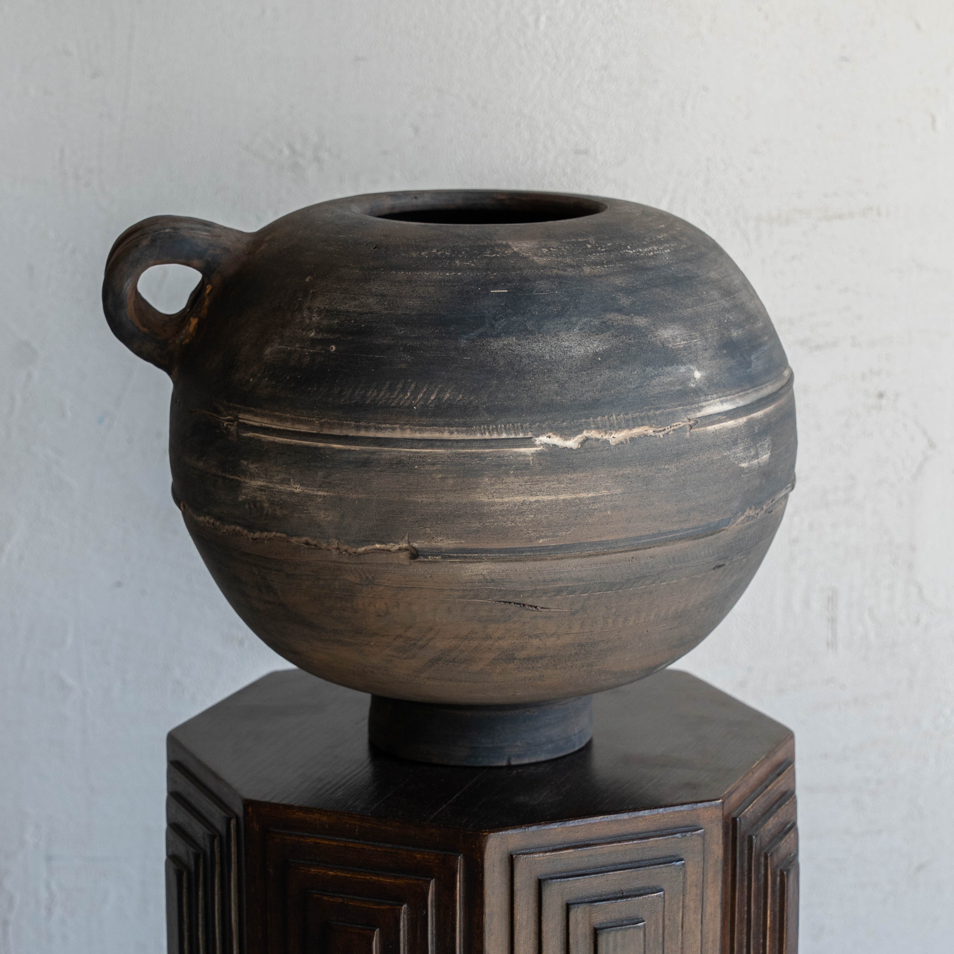 Dark brown ceramic vase on a hexagonal wooden stand against a light gray wall