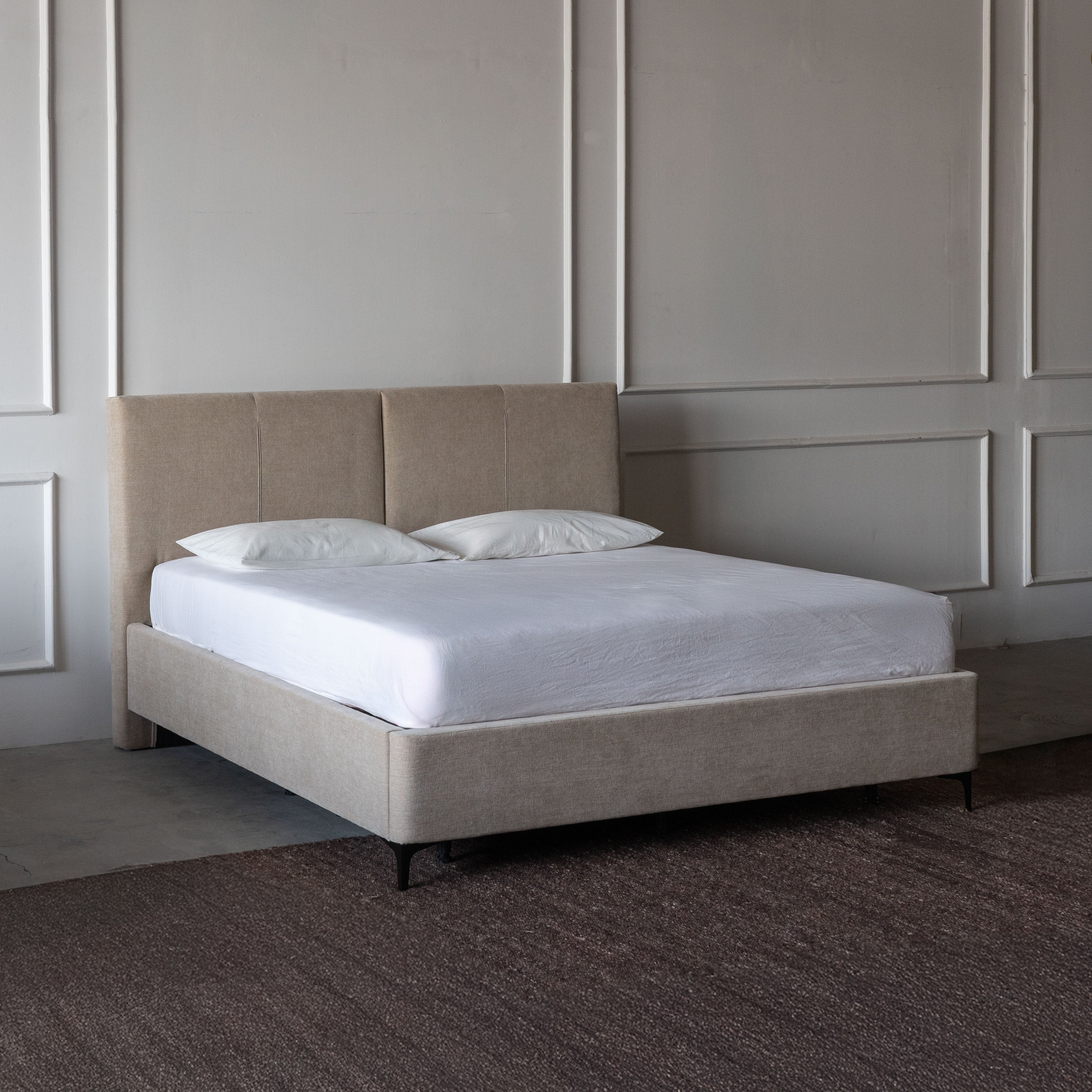 Beige upholstered bed with white bedding against a neutral wall.