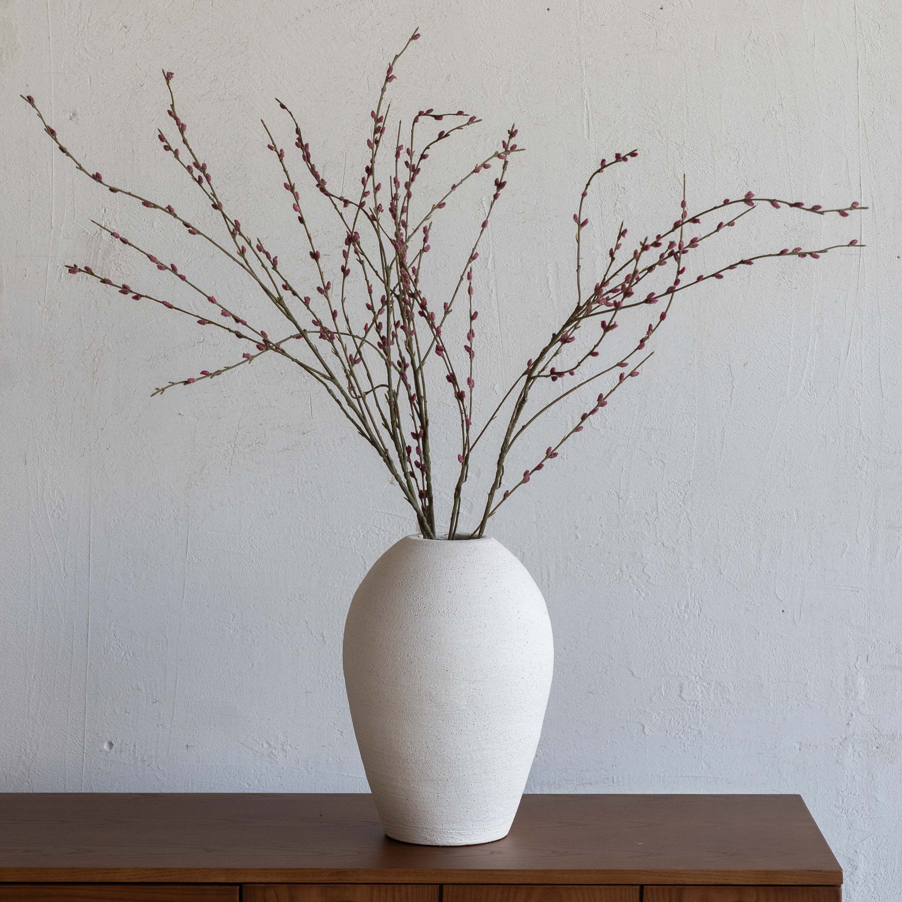 White vase with dried branches on a wooden cabinet against a plain wall
