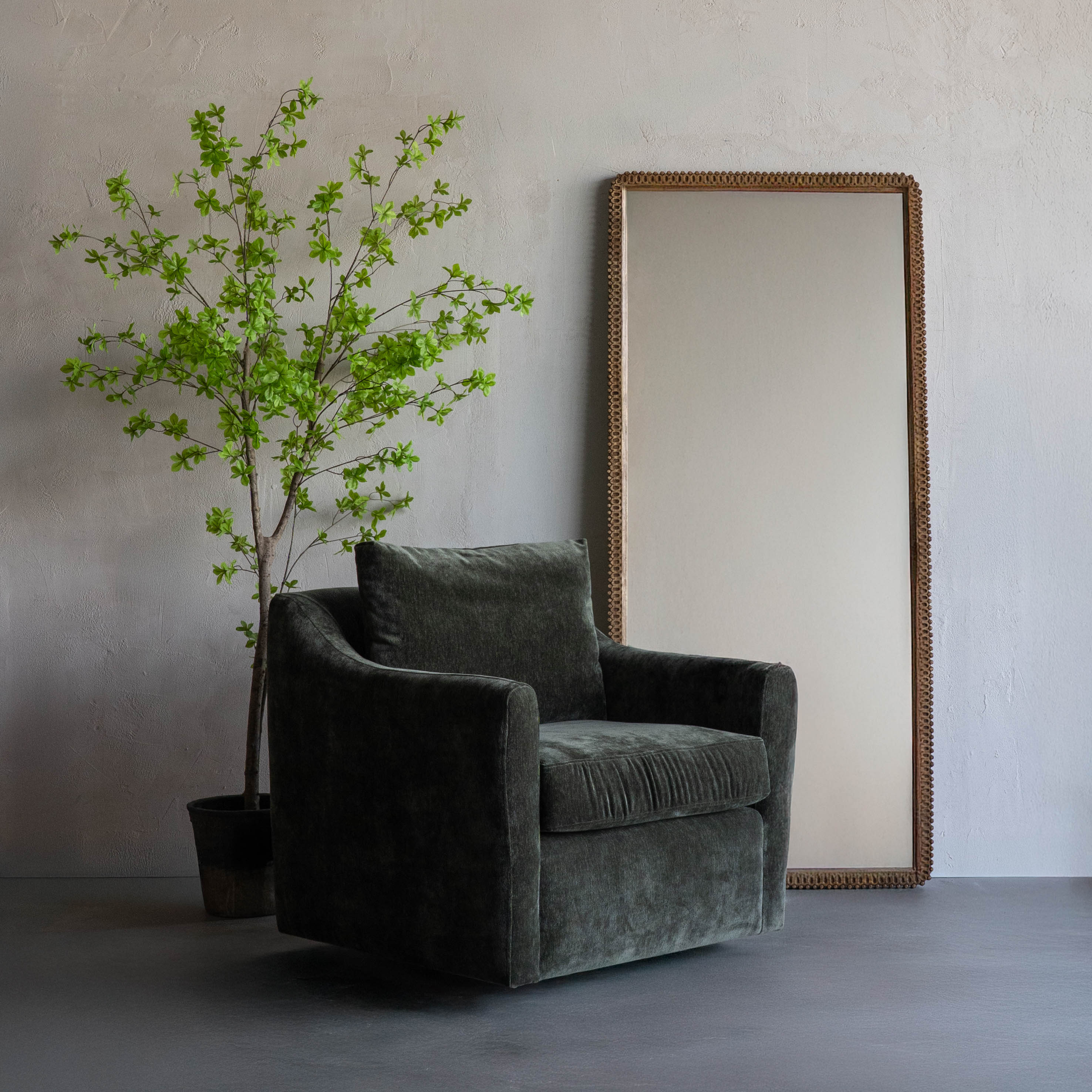 Gray armchair next to a potted plant and a large mirror on a plain wall.