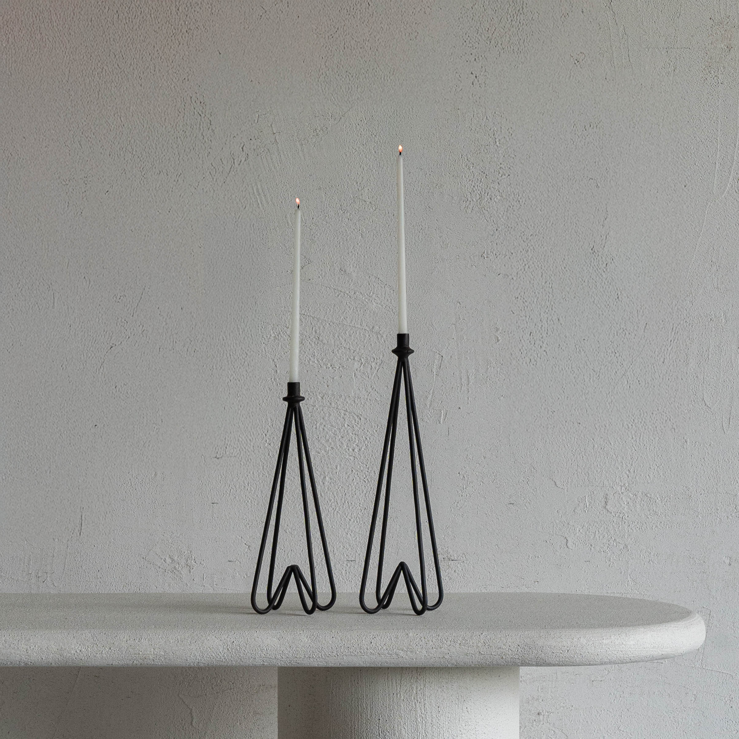 Two black wire candle holders with white candles on a gray surface against a gray wall.