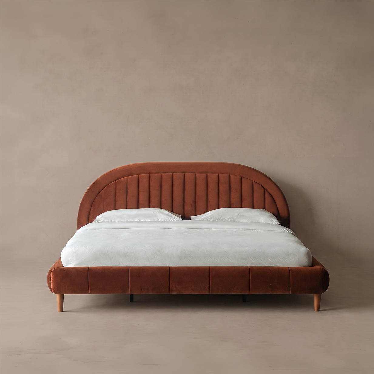 A modern platform bed with a curved, vertically channel-tufted headboard and matching footboard in a warm rust-brown velvet, styled with white bedding against a textured beige wall.