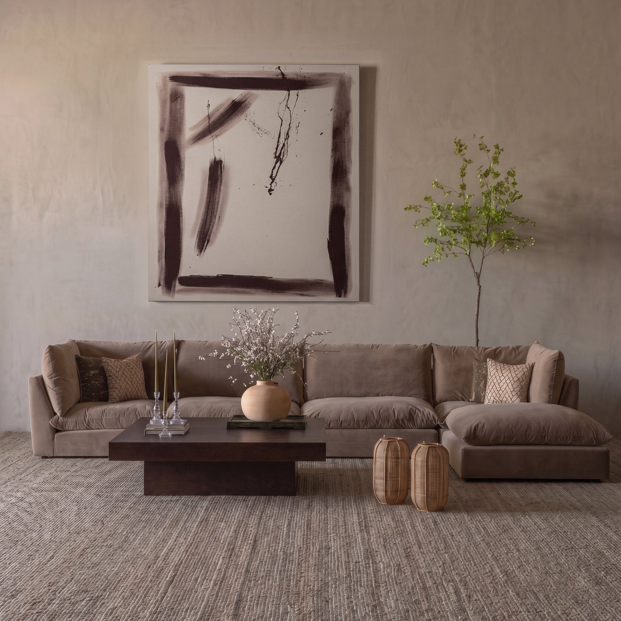 A staged living room scene featuring a large L-shaped sectional sofa upholstered in taupe fabric, set against a textured beige plaster wall with a large abstract painting. A dark wood coffee table, decorative vases, and a small potted tree complete the room.