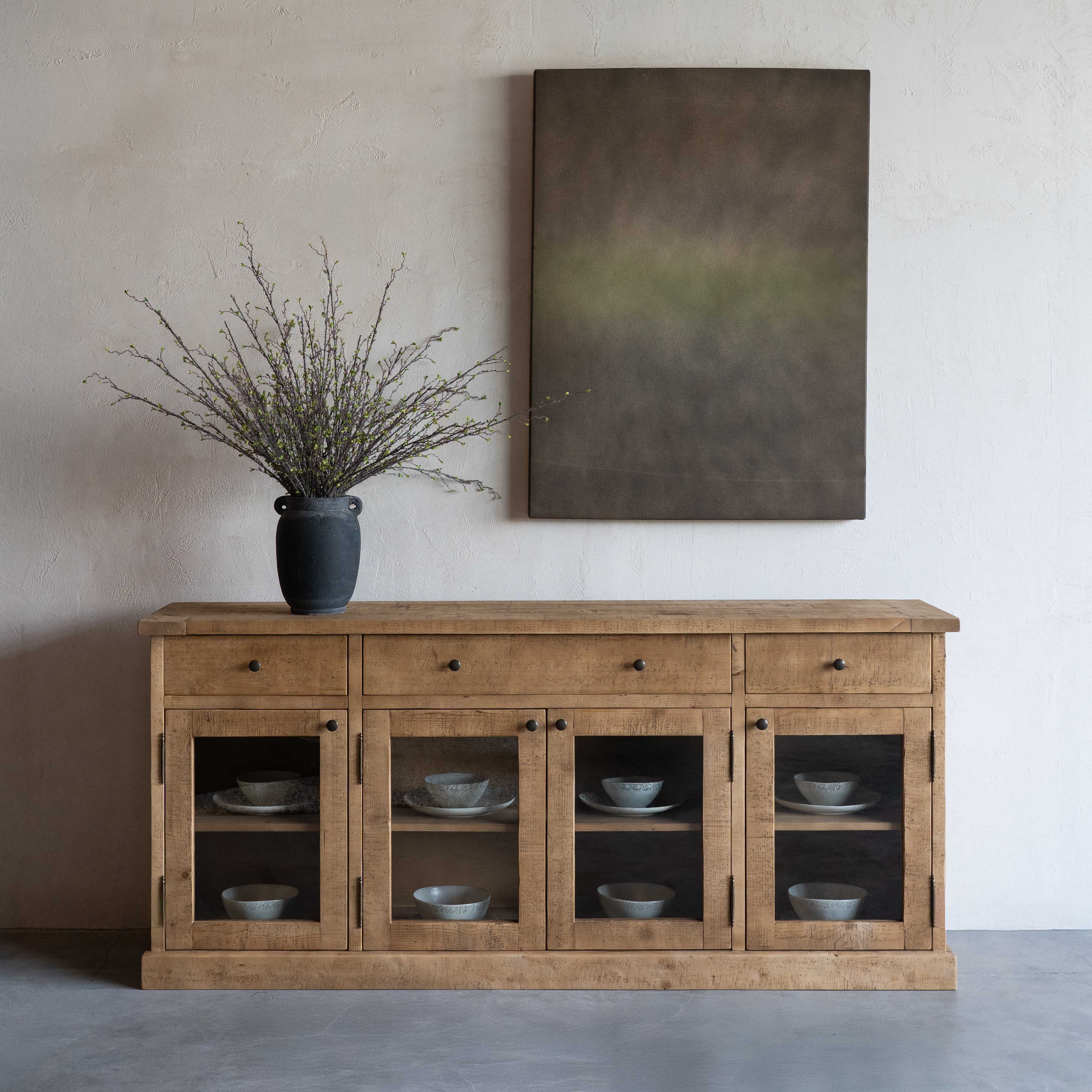 Wooden sideboard with glass doors displaying bowls, vase with branches on top, and abstract painting on wall