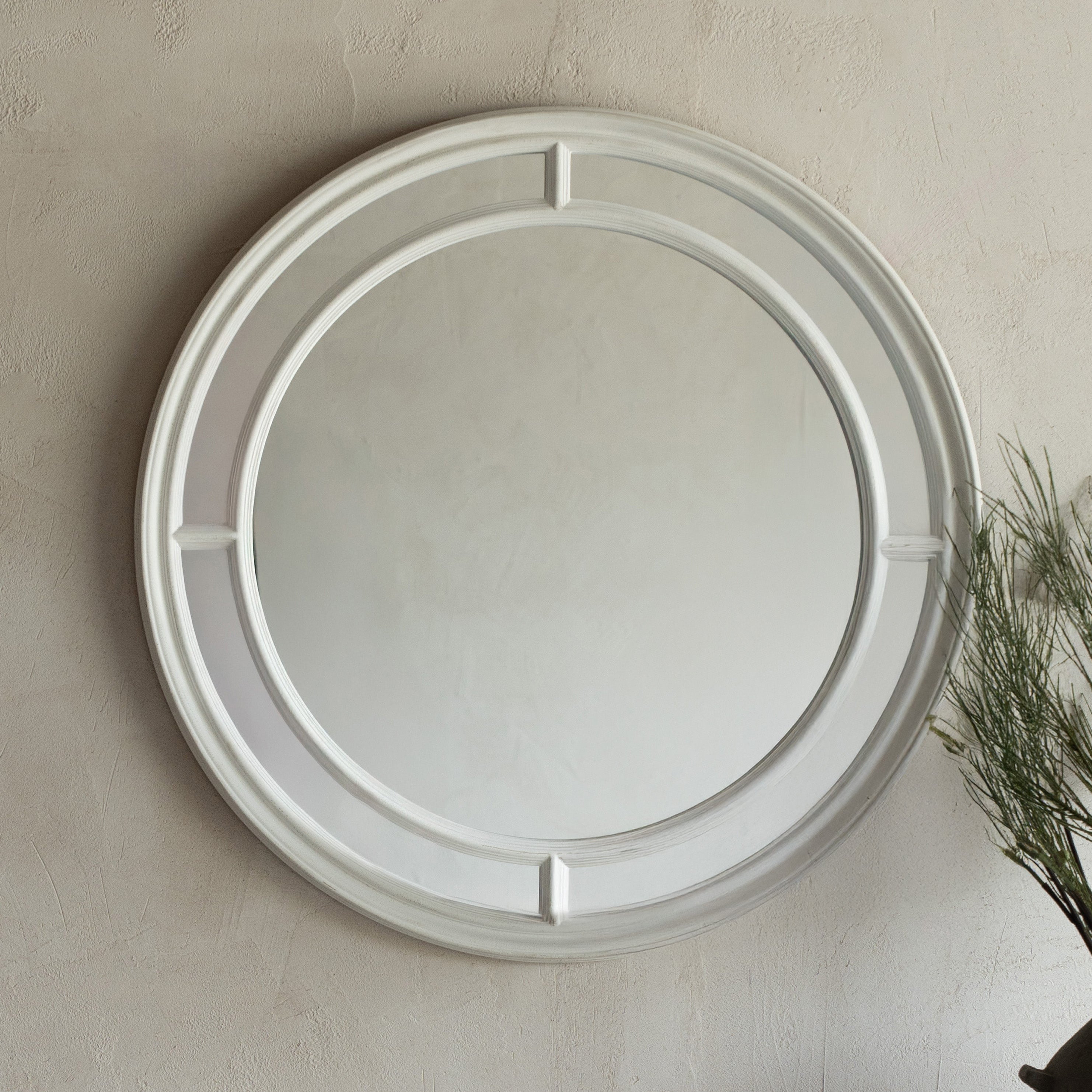 Round mirror with decorative frame on a beige wall above a white console table with a dark vase containing greenery.