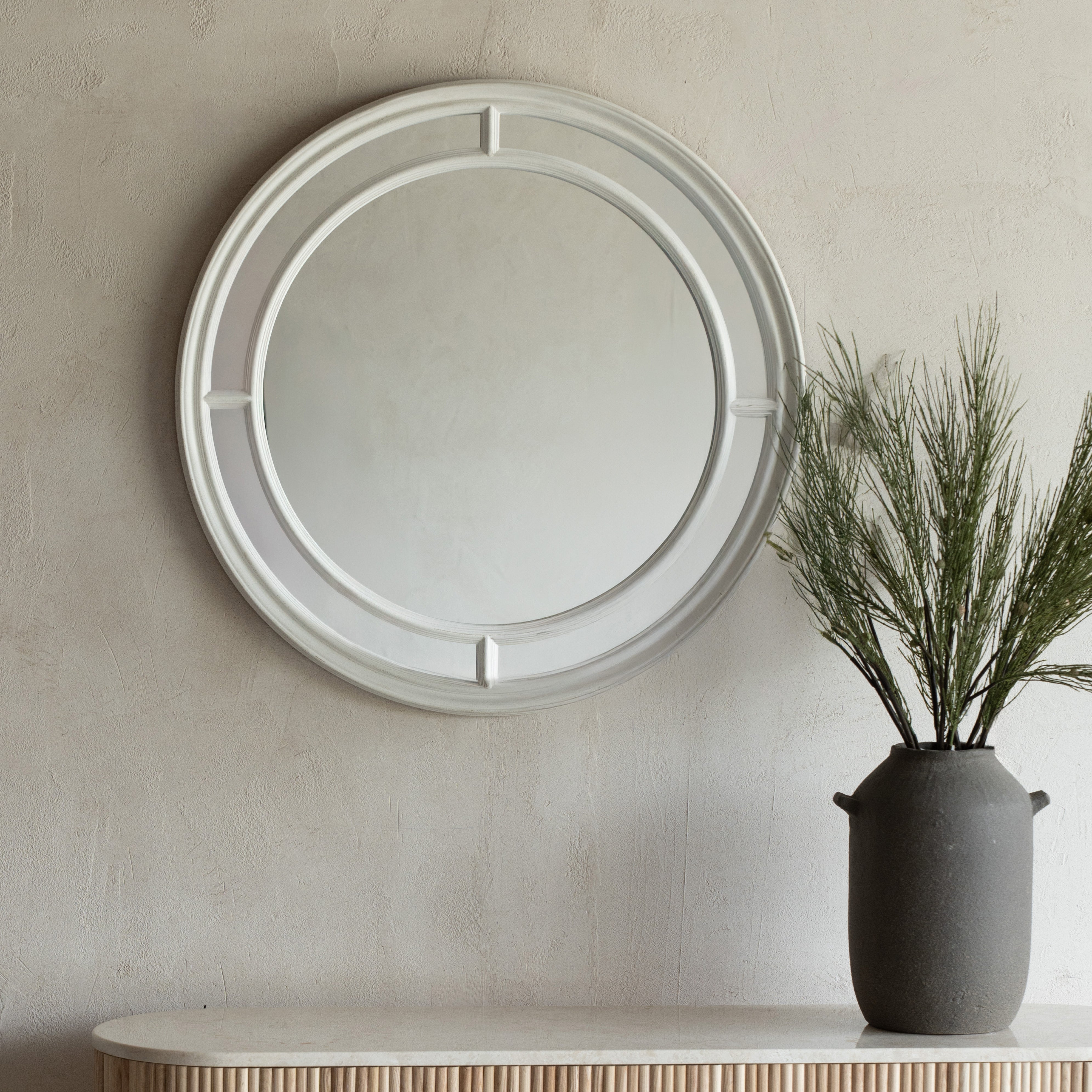 Round mirror with decorative frame on a beige wall above a white console table with a dark vase containing greenery.