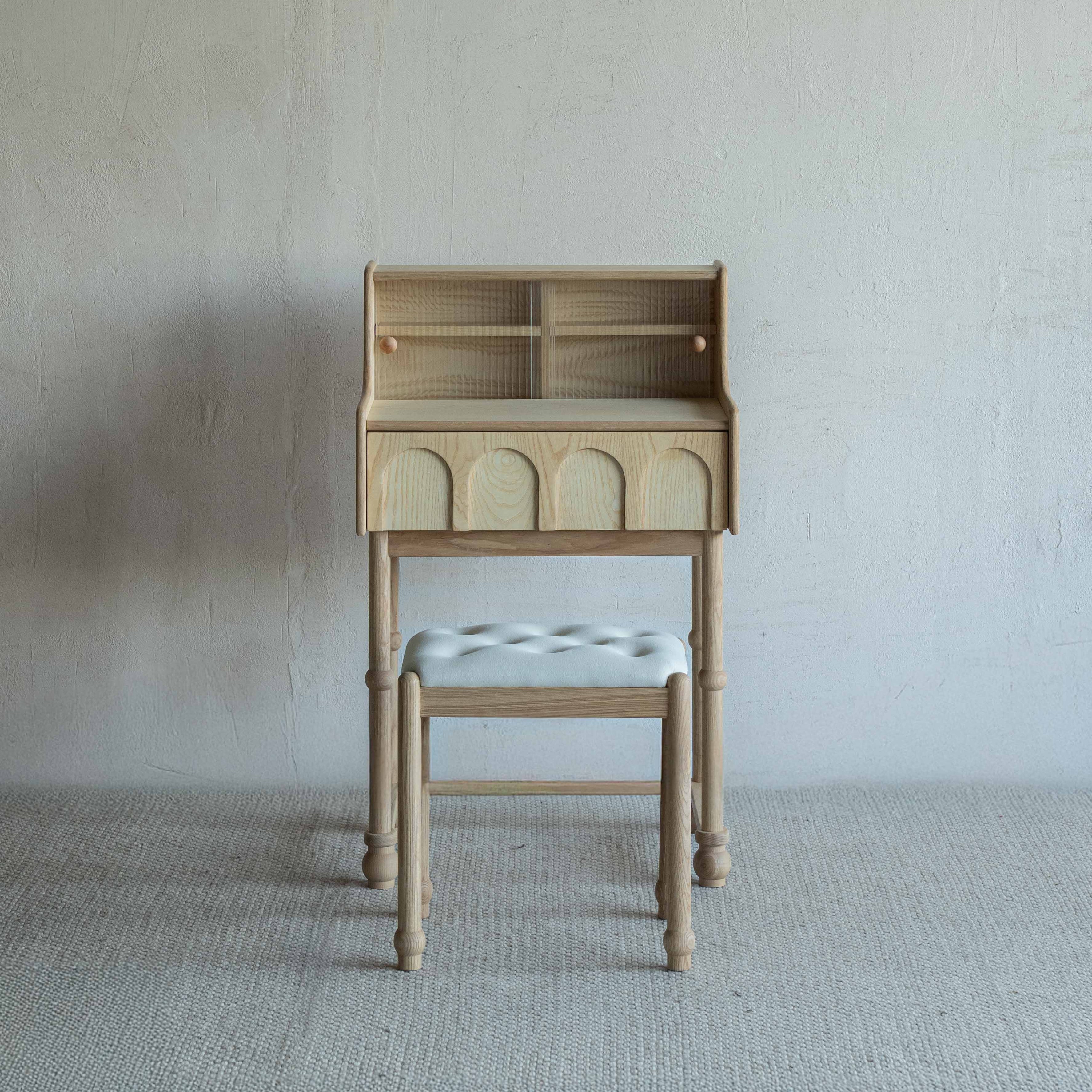 Wooden desk with a cushioned stool against a plain wall.